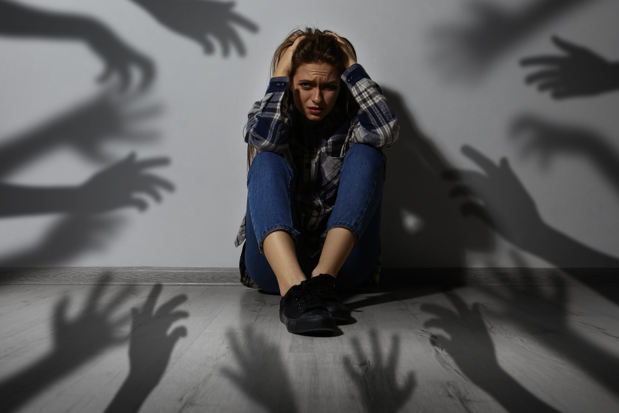 A distressed woman sits against a wall, clutching her head, as shadowy hands reach toward her from behind.
