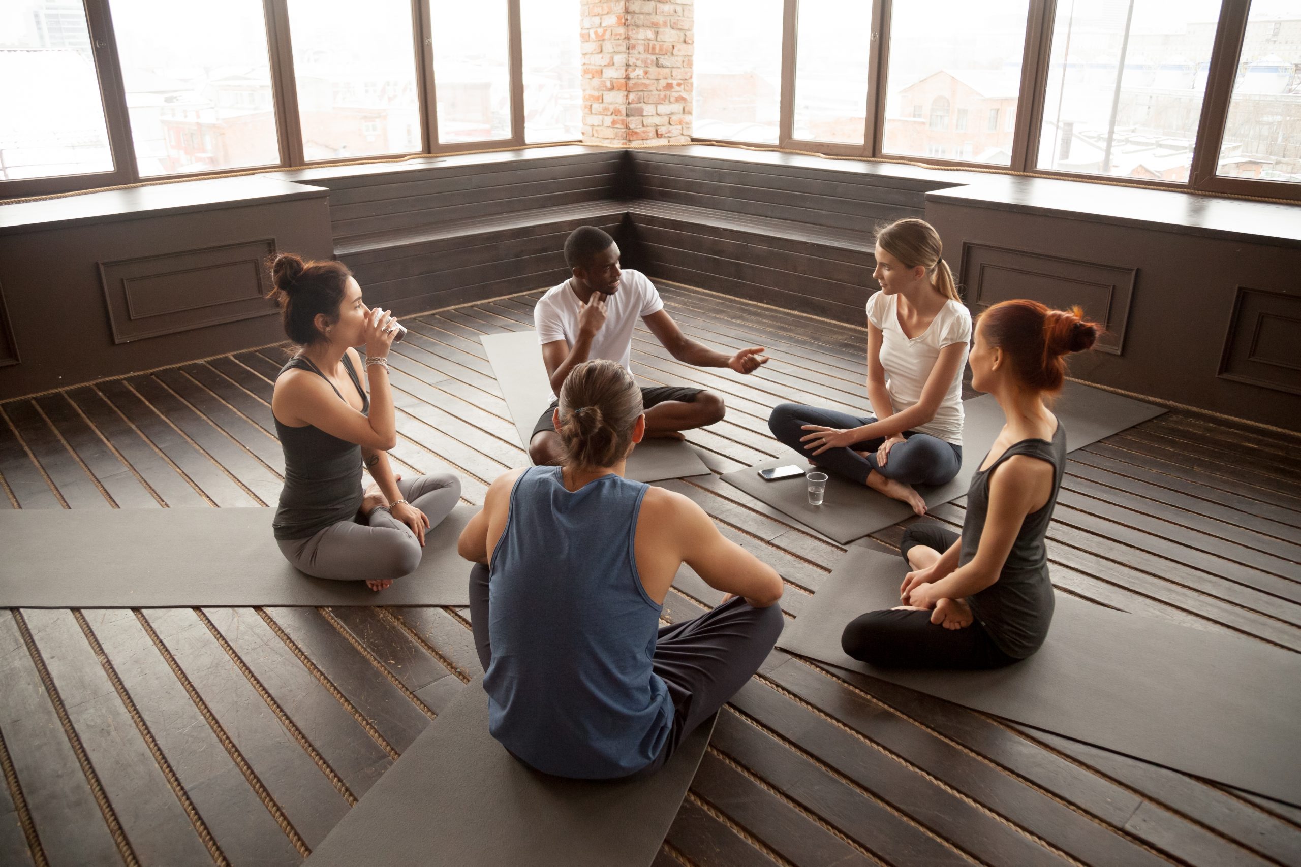 A diverse group of five people sitting on yoga mats, engaged in conversation in a bright, airy studio.