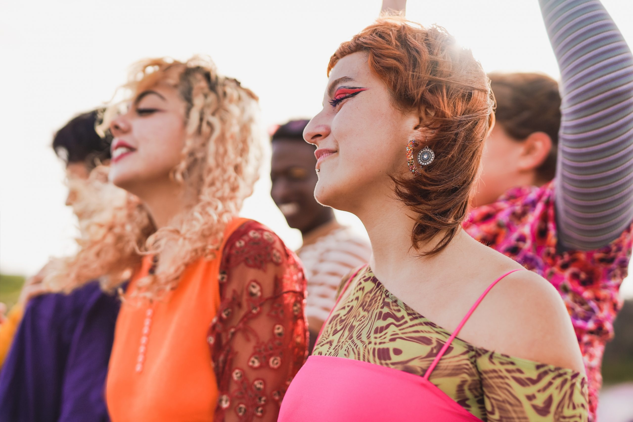 A diverse group of young women in vibrant outfits, enjoying a lively moment outdoors, with colorful hairstyles and makeup.