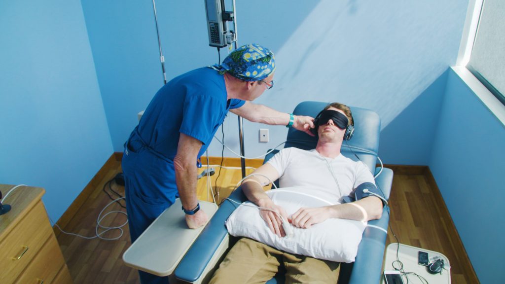 A doctor in scrubs adjusts a blindfolded patient lying on a treatment chair in a bright, clinical room.