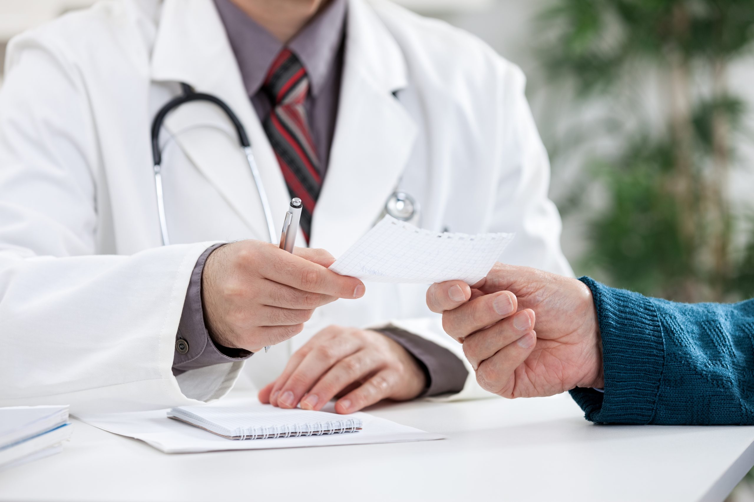 A doctor in a white coat hands a prescription note to a patient in a blue sweater.