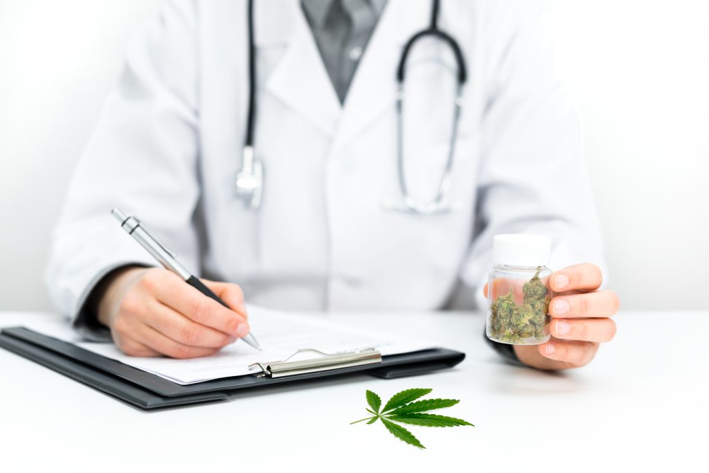 A doctor in a white coat holds a jar of cannabis while writing notes on a clipboard. A cannabis leaf is on the table.
