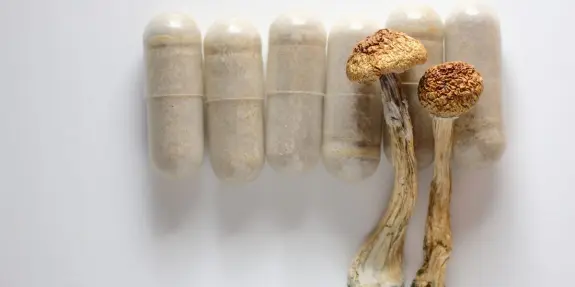 Dried psilocybin mushrooms beside several clear capsules filled with a light brown powder.