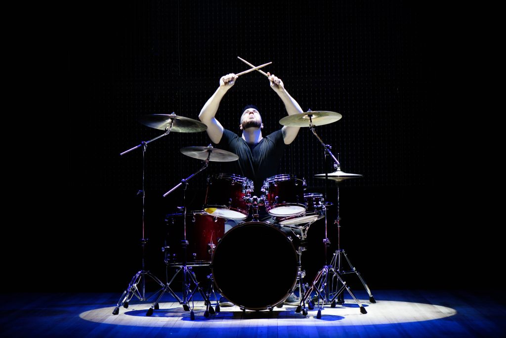 Drummer passionately performing on stage, illuminated by spotlight, with drumsticks raised above a vibrant drum set.