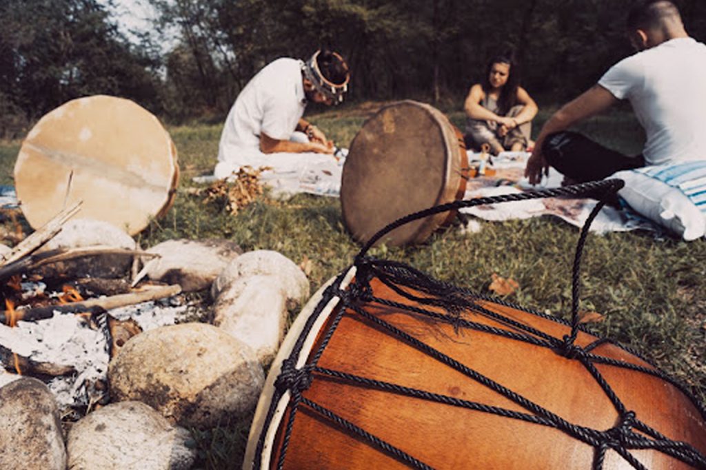 Drummers gather around a fire, crafting instruments and sharing a cultural experience in a serene outdoor setting.