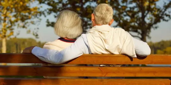 Elderly couple sitting on a bench, embracing, surrounded by trees with autumn leaves.