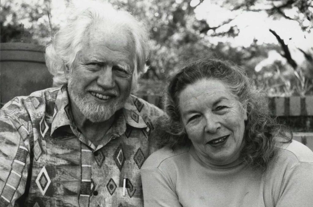 Elderly couple smiling together, with a blurred natural background and patterned clothing.