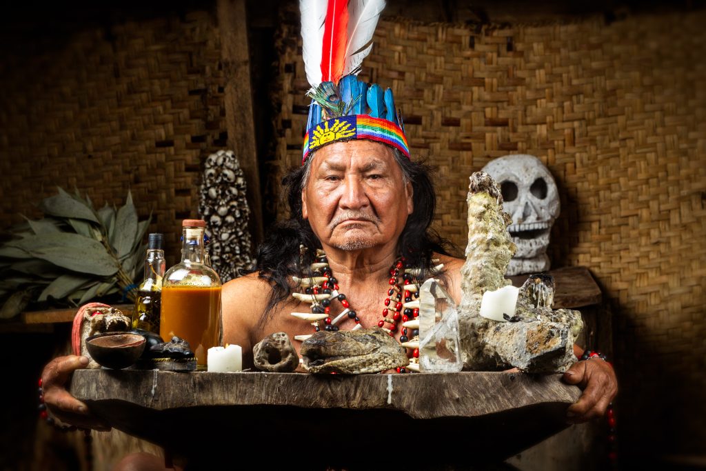 Elderly Indigenous man in traditional attire holds a wooden tray with bottles, stones, and candles, surrounded by natural ...