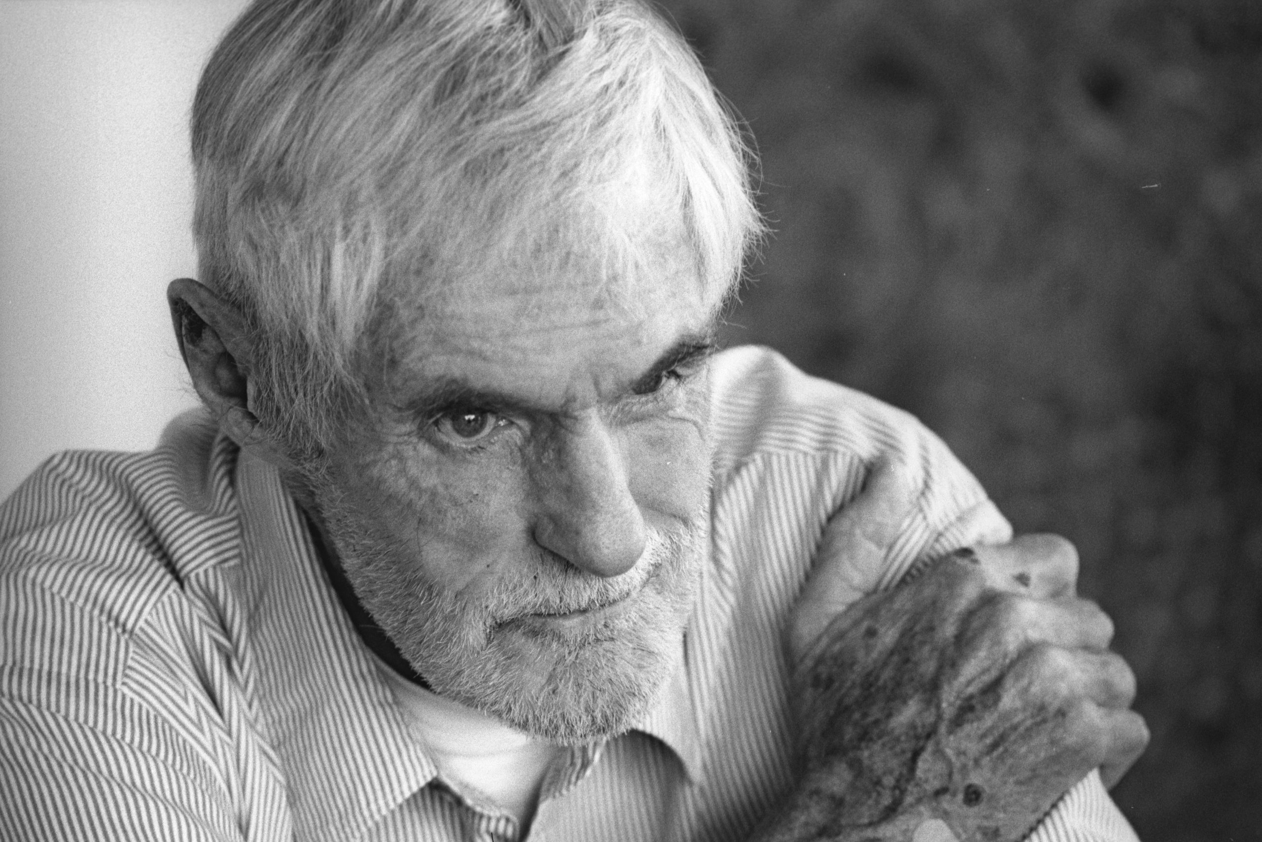 Elderly man with gray hair and beard, deep in thought, resting his chin on crossed arms, in soft focus background.