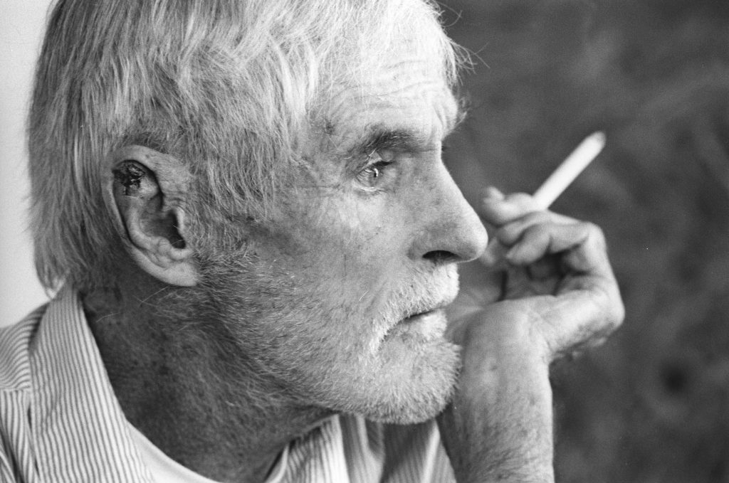 Elderly man with gray hair and a beard, holding a cigarette, gazing thoughtfully to the side.