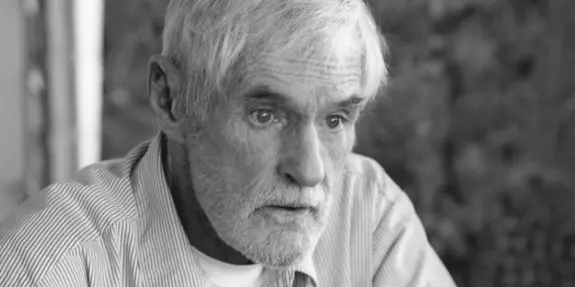 Elderly man with gray hair and a thoughtful expression, wearing a striped shirt against a blurred background.