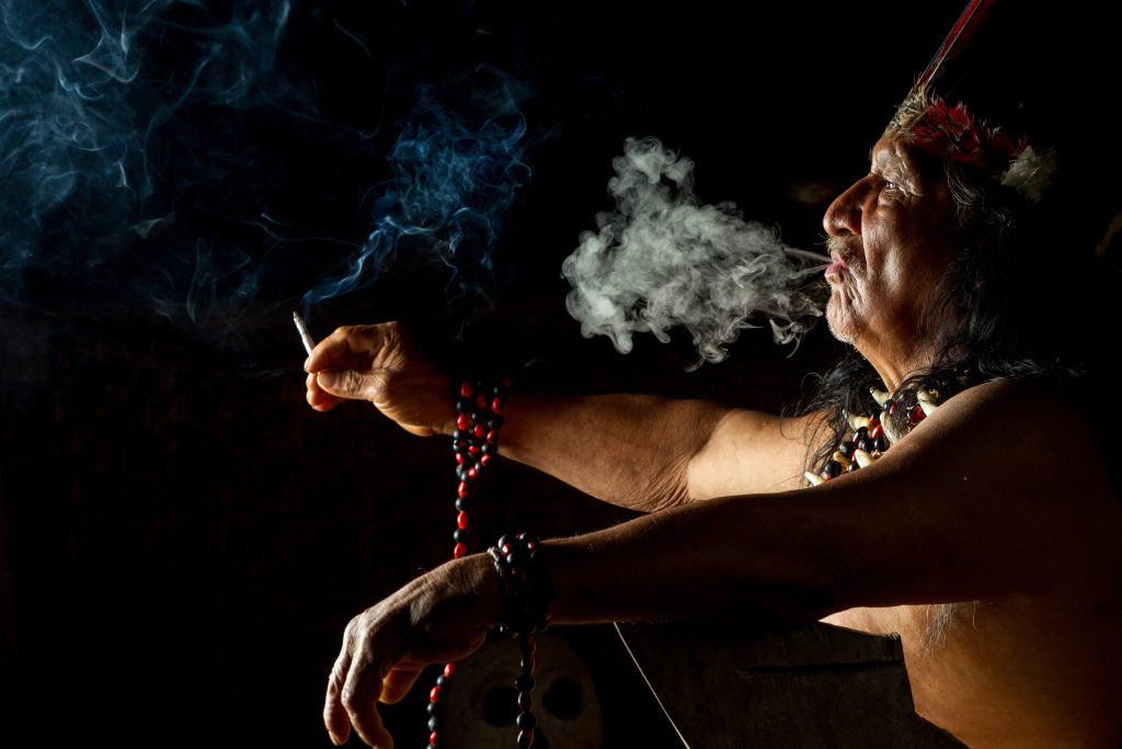 Elderly man with long hair and traditional attire exhales smoke, adorned with beads and a feathered headdress.