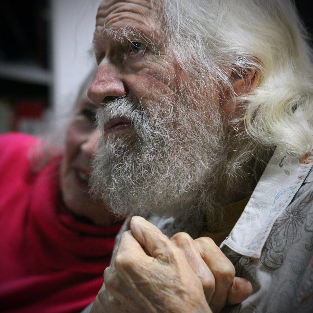 Elderly man with long white hair and a beard, holding hands, deep in thought, with a woman softly smiling in the background.