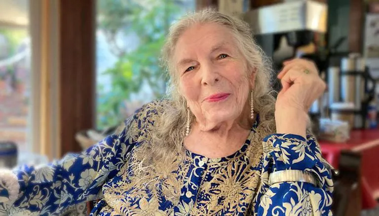 Elderly woman with long gray hair, wearing a blue and gold patterned top, smiling warmly in a cozy indoor setting.