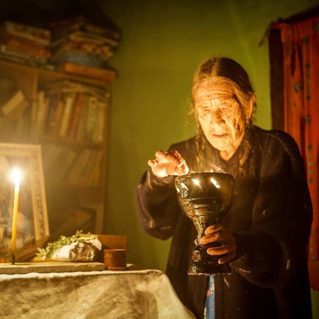 Elderly woman with long gray hair holds a glowing chalice, surrounded by candlelight and a cozy, cluttered room.