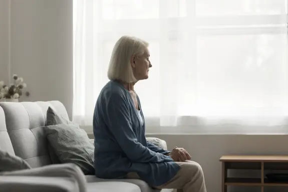 Elderly woman with short gray hair sits pensively on a couch, gazing out a window with soft natural light.