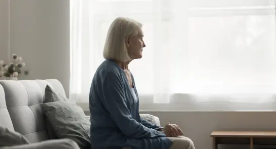 Elderly woman with short gray hair sits pensively on a couch, gazing out a window with soft natural light.