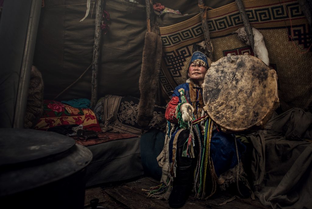 Elderly woman in traditional attire plays a drum inside a cozy, decorated tent with fur and patterned textiles.