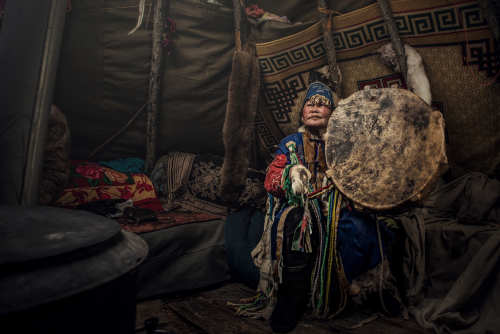 Elderly woman in traditional attire plays a drum inside a cozy tent adorned with patterned textiles and fur.