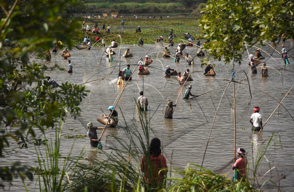 Fishermen wade through water, casting nets among lush greenery, with a bustling scene of activity in the background.