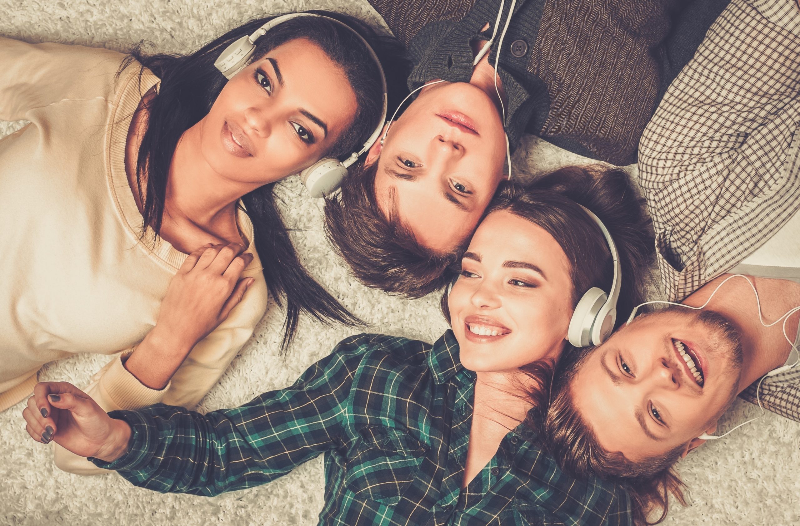 Four friends lie on a soft surface, wearing headphones and smiling, enjoying music together in a cozy setting.