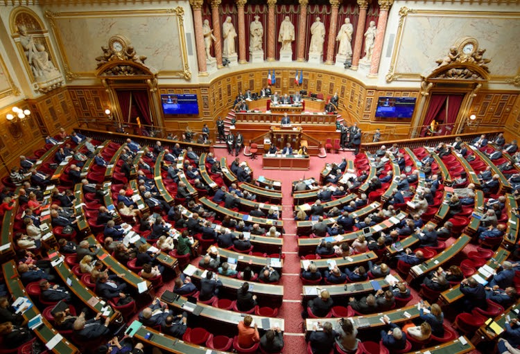 French Senate chamber filled with seated senators, ornate decor, and a central podium for speeches.