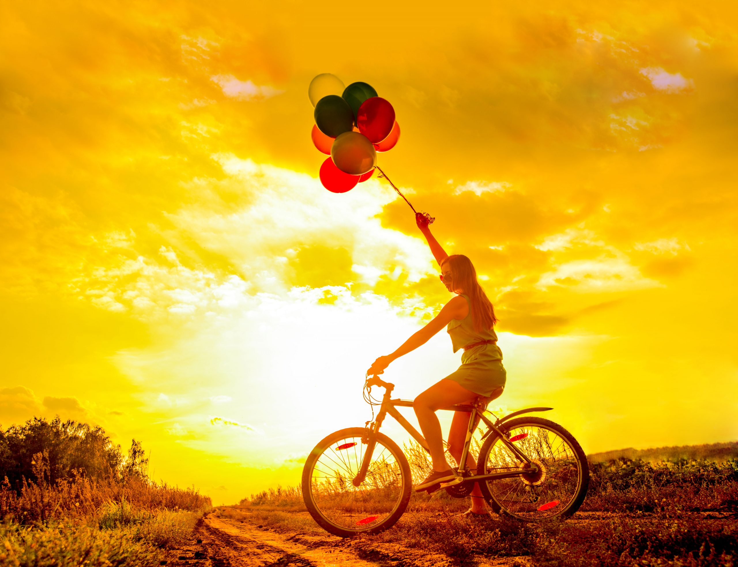 A girl on a bicycle holds colorful balloons against a vibrant sunset sky, creating a joyful and carefree atmosphere.