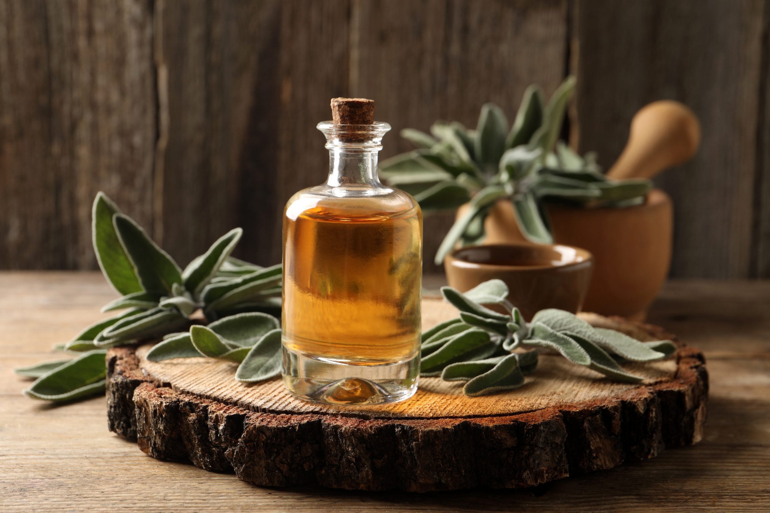 Glass bottle of golden oil on a wooden slice, surrounded by fresh sage leaves and a wooden mortar and bowl.