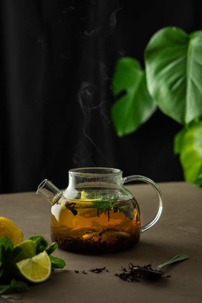 Glass teapot with steaming herbal tea, mint leaves, and lemon slices, set against a dark background with green foliage.