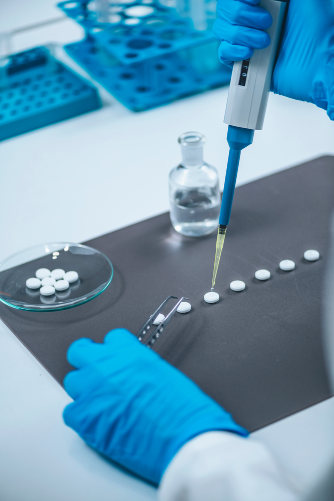 A gloved hand uses a pipette to dispense liquid onto white tablets on a lab surface, with glassware in the background.