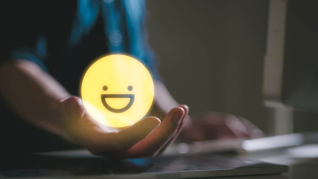 A glowing yellow smiley face held in a hand, with a blurred background of a computer setup.