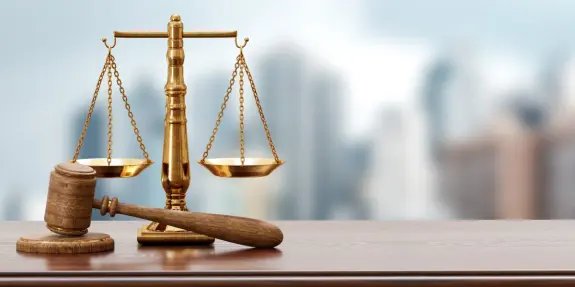 Golden scales of justice beside a wooden gavel on a polished table, with a blurred cityscape in the background.
