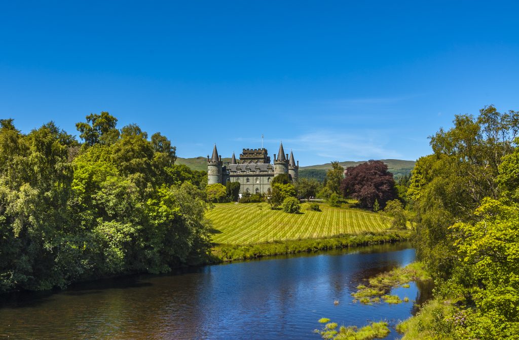 A grand castle surrounded by lush greenery and a serene river under a clear blue sky.