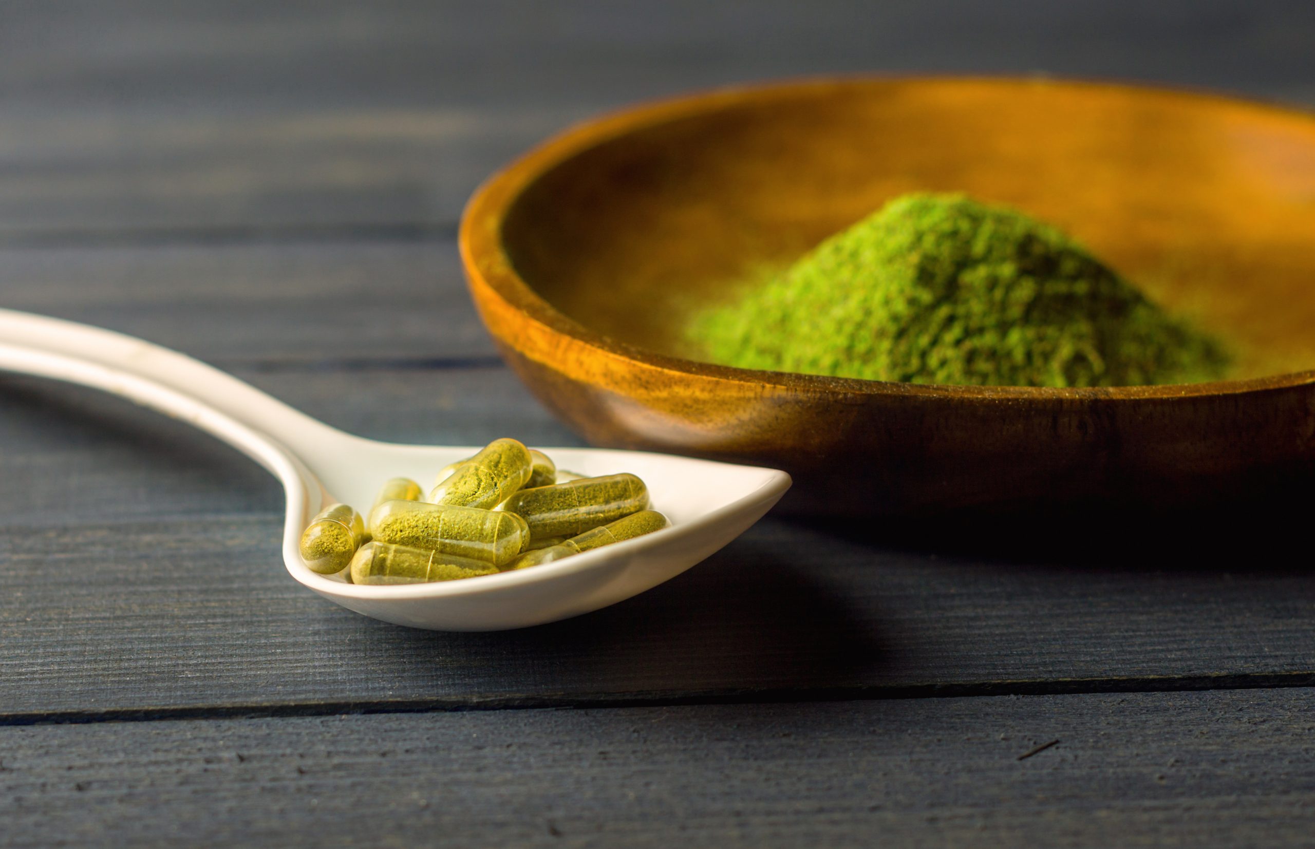 Green capsules on a white spoon beside a wooden bowl filled with green powder, set against a dark wooden background.