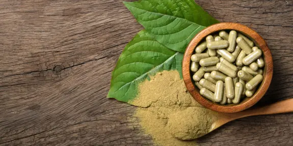Green capsules in a wooden bowl, surrounded by green leaves and a pile of powder on a rustic wooden surface.