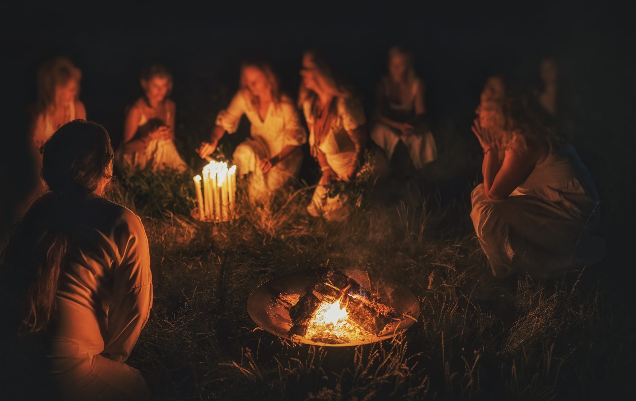 A group of women in white attire gathered around a glowing campfire at night, sharing stories and laughter.