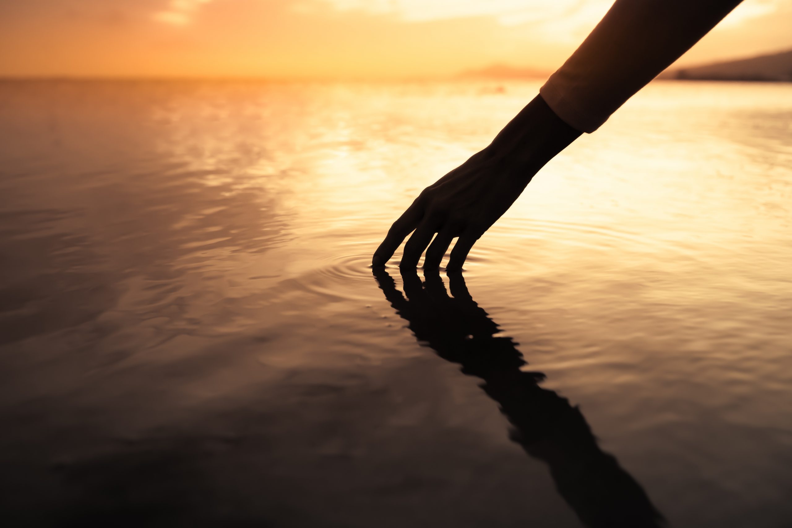 A hand gently touches the water's surface at sunset, creating ripples against a golden sky.