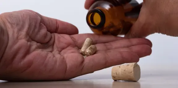 Hand holding capsules as another hand pours them from a brown bottle, with a cork lying nearby.