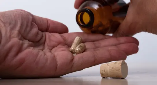 Hand holding capsules as another hand pours them from a brown bottle, with a cork lying nearby.