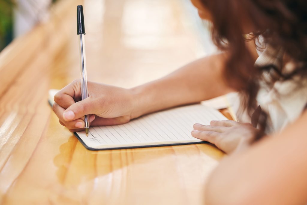 A hand holding a pen writes in a lined notebook on a wooden surface, with soft natural light illuminating the scene.