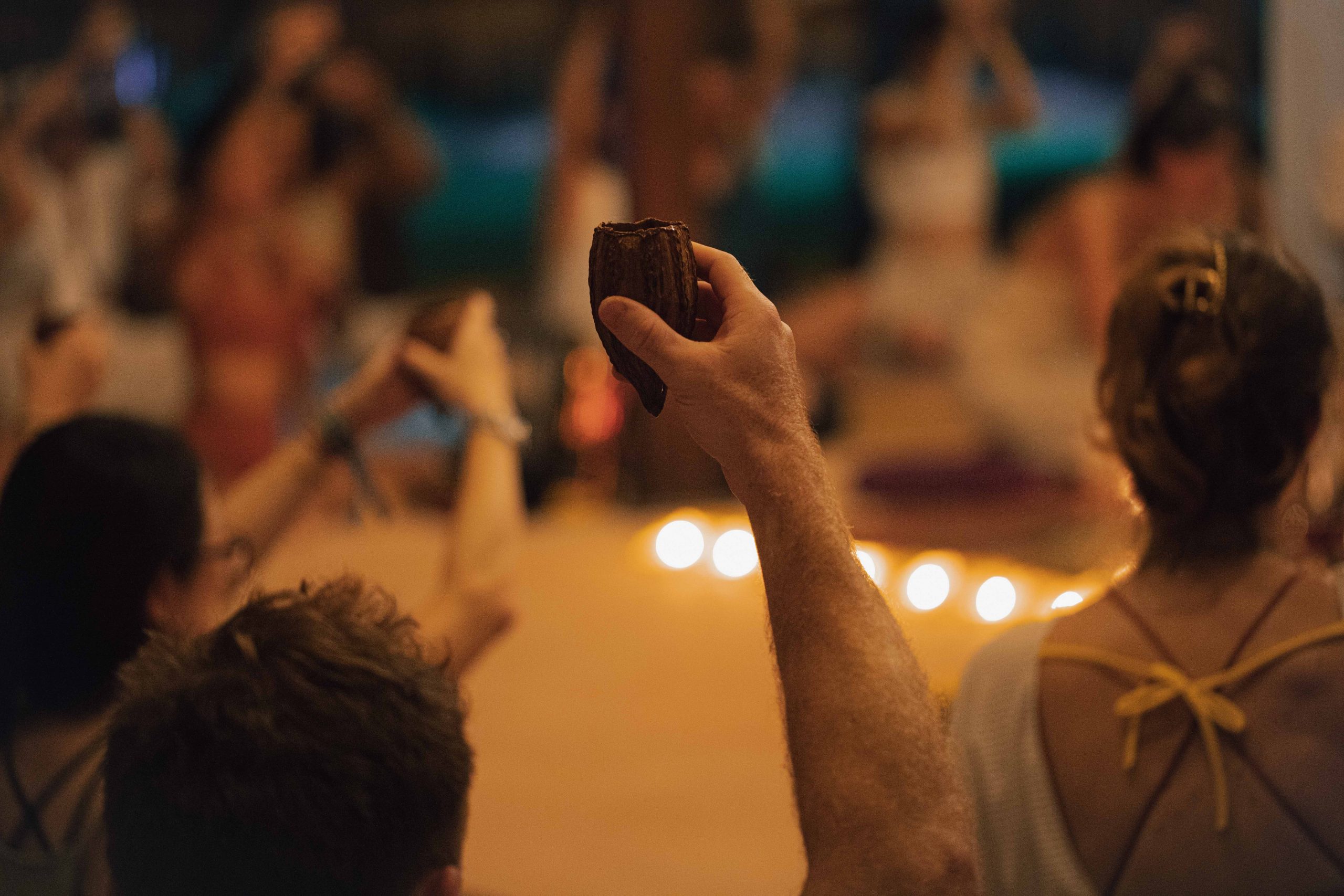 A hand holds a ceremonial drink, surrounded by participants in a dimly lit gathering with glowing lights.