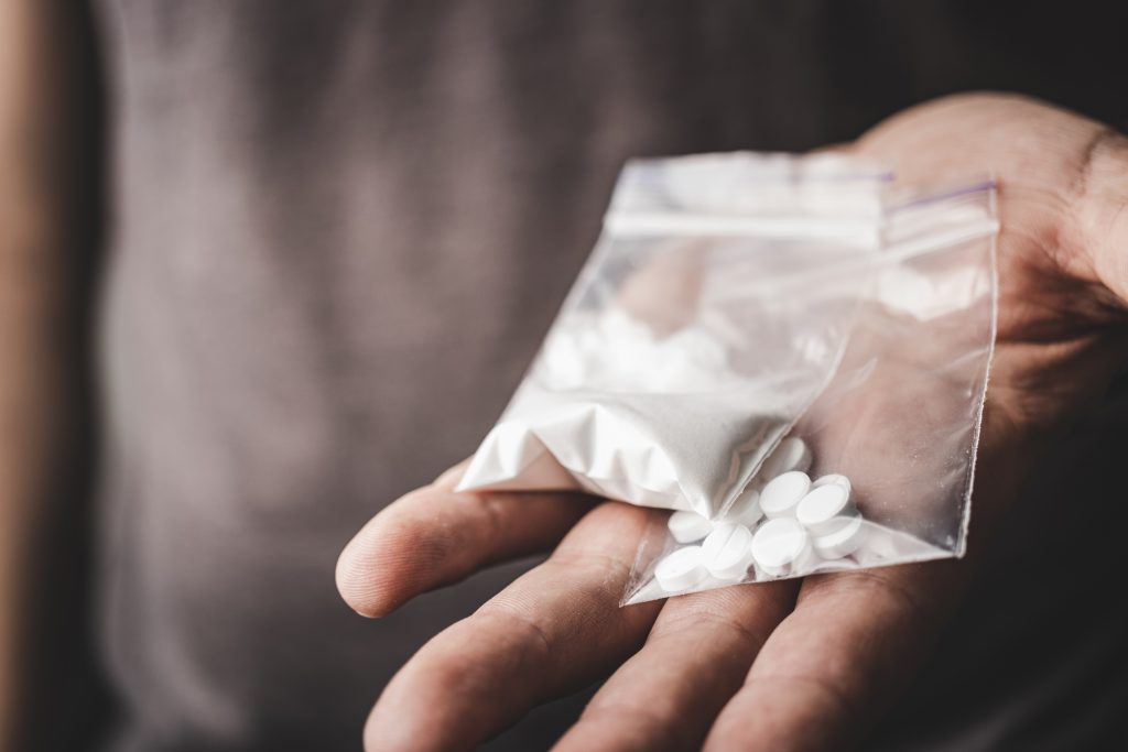 A hand holds a clear bag of white powder alongside several white pills.
