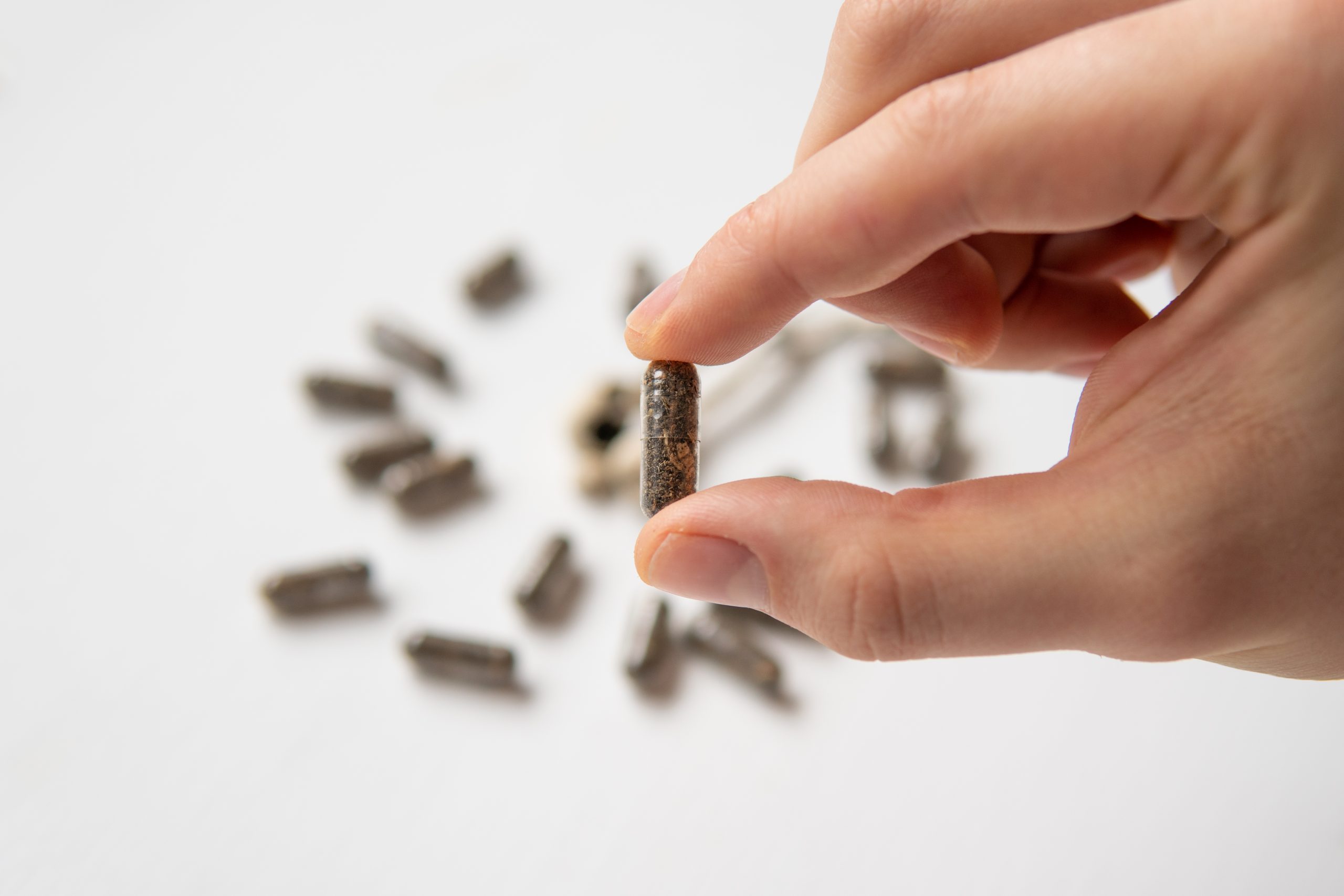 A hand holds a single brown capsule above scattered capsules on a white surface.