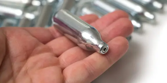 A hand holds a small, shiny silver canister among several similar canisters in the background.