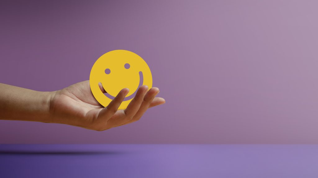 A hand holds a yellow smiley face against a soft purple background.