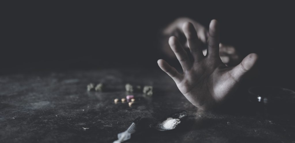 A hand reaches out from a dark surface, surrounded by scattered pills and a small pile of white powder.