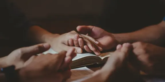 Hands clasped together in prayer over an open book, symbolizing unity and reflection. Soft, warm lighting enhances the scene.