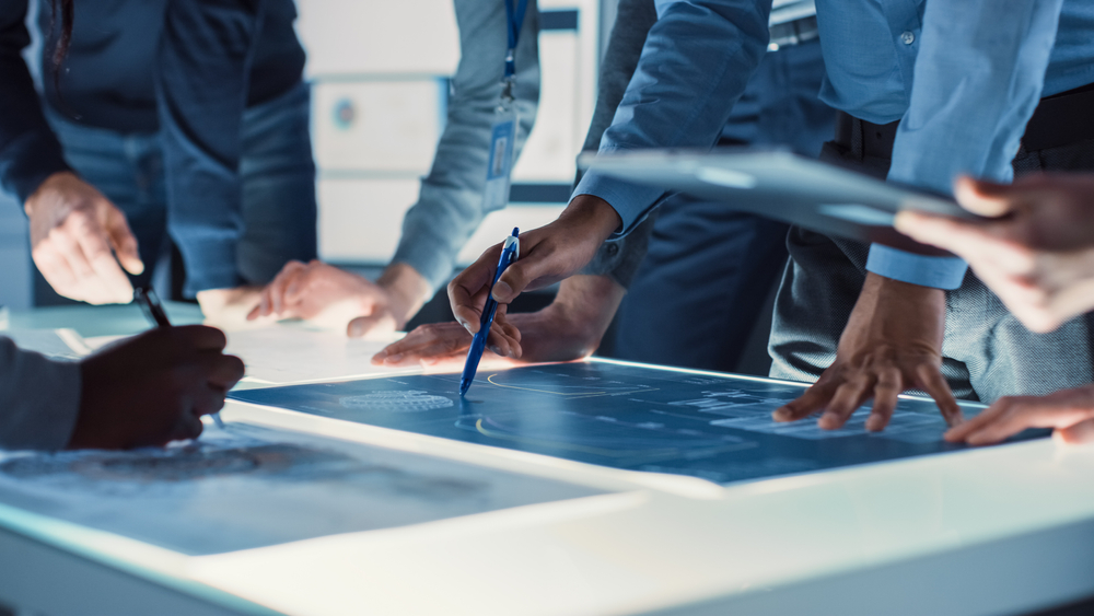 Hands of diverse individuals collaborating over illuminated blueprints on a table, with pens and tablets in use.