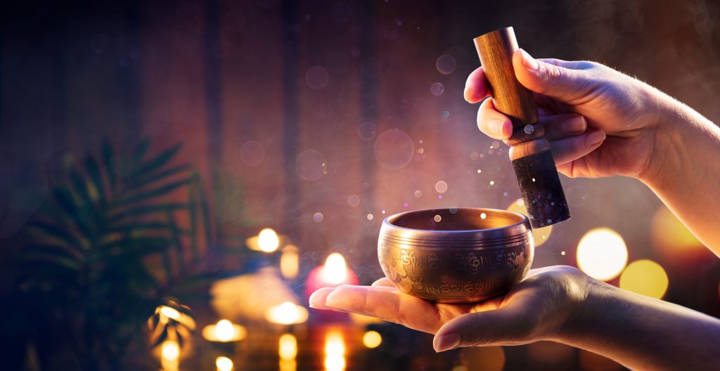 Hands gently strike a bronze singing bowl, releasing soft vibrations amidst a serene, candlelit atmosphere.