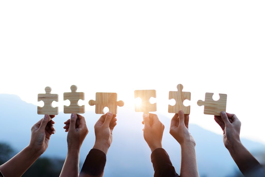 Hands hold wooden puzzle pieces against a bright sky, symbolizing unity and collaboration.